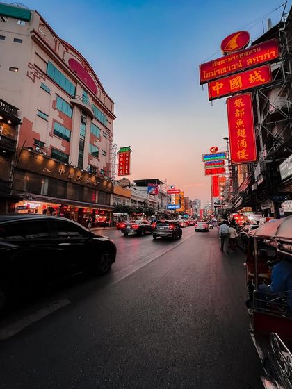 The view of a Bangkok street at dusk, with cars and tuk-tuks making their way through the city under the glow of Chinese signs.