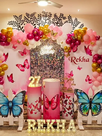 An elegant butterfly theme for a 27th birthday, perfect for adults. The design uses a pink and gold balloon arch over a silver shimmer wall, accented with large butterfly cutouts and marquee letters for the name.