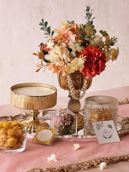 The Bright Bliss hamper is designed to delight. It features a large candle, a box of truffles, a glass jar with butter cookies, and a flower arrangement on a mirror tray.