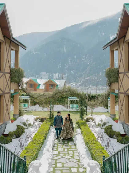 A stunning wide shot of the couple walking in a manicured garden with snow and mountains in the background, capturing the magic of their Manali Mehendi.