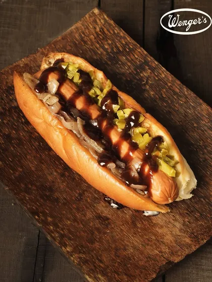 Our take on the classic Hot Dog. This image shows a juicy sausage nestled in a soft, fresh bun from our bakery, topped with onions, relish, and a generous drizzle of sauce.
