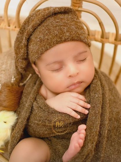 A close-up of a sleeping newborn in a brown knit outfit, nestled in a small wicker chair with a tiny chick prop.