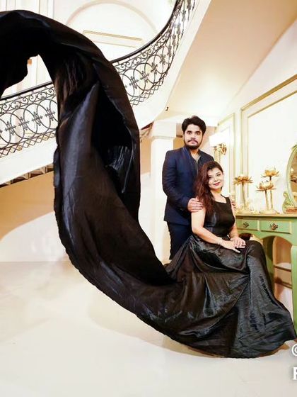 A couple poses in a grand hall, the bride seated while her black trail gown flows dynamically, creating an artistic and modern pre-wedding photo.