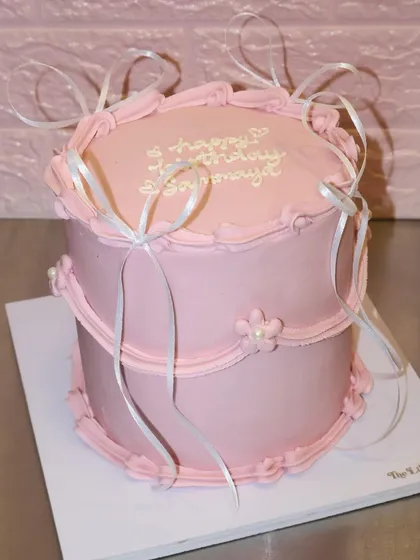 Turning one never looked so cute. This elegant two-tiered pink cake is decorated with simple piping and delicate white ribbons, perfect for a first birthday.