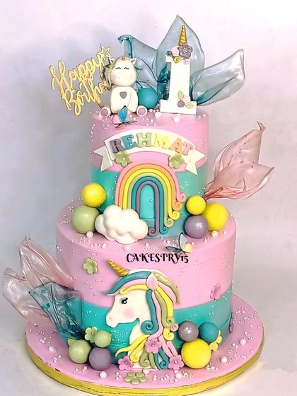 An enchanting two-tier unicorn-themed cake. The design features a mix of pink and aqua colors, a large fondant unicorn, a rainbow, and delicate rice paper sails.