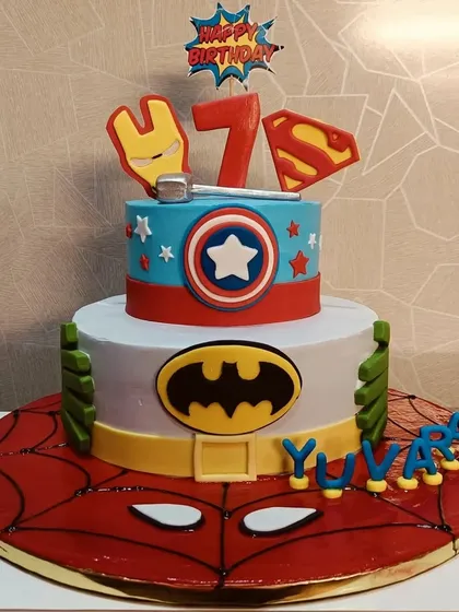 A two-tiered superhero cake combining the worlds of Marvel and DC. It features logos and elements from Batman, Superman, Captain America, and Iron Man, all on a Spider-Man base.