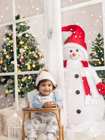A happy baby enjoying the magical snowfall from a cozy chair, with our friendly snowman peeking through the window.