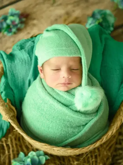 A perfect little bundle. This classic newborn pose shows a baby swaddled in a soft green wrap, nestled peacefully in a rustic basket. The pom-pom hat adds a touch of playful charm.