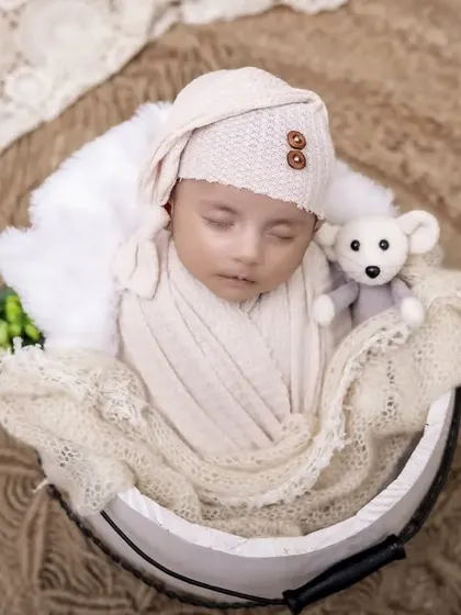 Dressed as a little angel with tiny wings, this newborn sleeps with a teddy bear. The neutral tones of the swaddle, hat, and basket create a soft and timeless look.