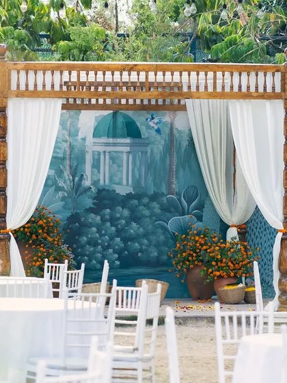 A simple yet beautiful mandap for a pre-wedding ceremony, with a hand-painted backdrop and elegant white seating.