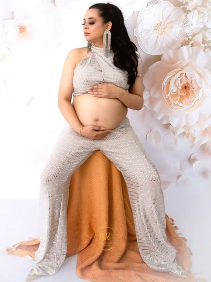 A modern and chic solo maternity portrait. The mother-to-be is seated, wearing a stylish silver sequined two-piece outfit against a white floral backdrop.