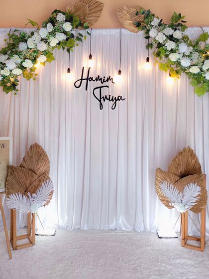A minimalist and modern wedding backdrop. This elegant setup features a clean white curtain, simple hanging bulbs, and delicate white floral arrangements, perfect for a chic and contemporary ceremony.