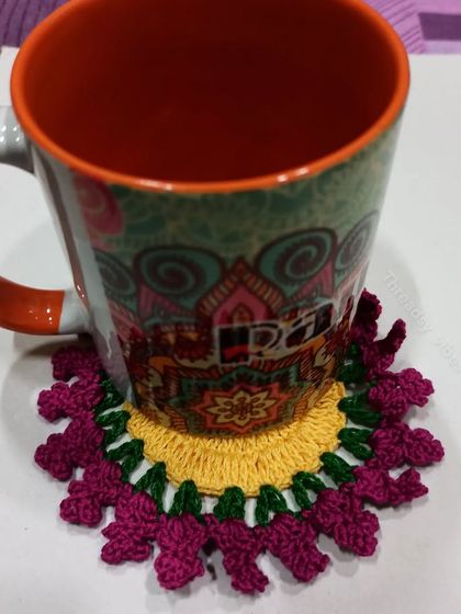 This floral crochet coaster uses a fun color combination of yellow, green, and magenta. It's pictured here with a mug, showing how it protects your surfaces in style.