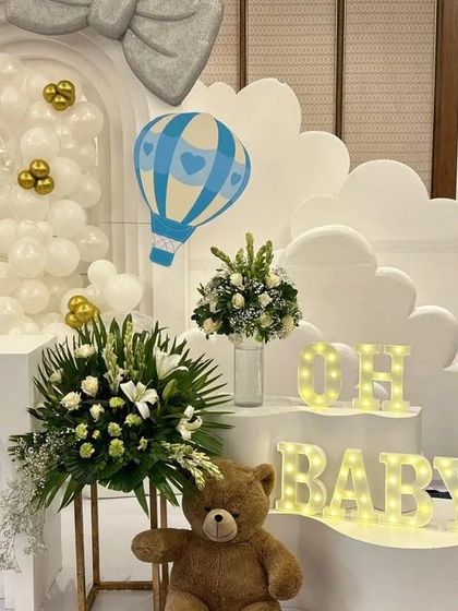 An elegant 'Oh Baby' setup with a hot-air balloon theme. The clean white structures, cloud cutouts, and marquee letters are accented with fresh flowers and a teddy bear for a classic and timeless look.