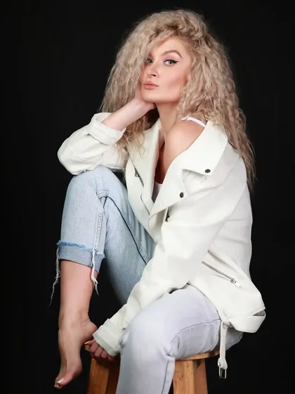 A classic portrait of a model with curly blonde hair in a white jacket, seated on a stool against a black background.