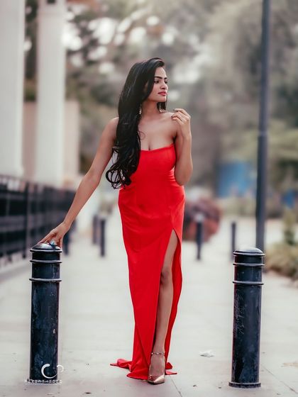 A full-length fashion shot on a city sidewalk. The flowing red dress with a high slit creates a glamorous and eye-catching silhouette.