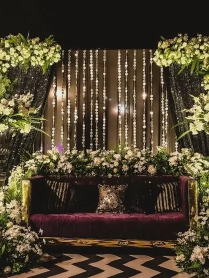 An opulent seating area for the couple at a reception. The rich purple sofa is framed by massive white floral arrangements and a glittering backdrop, creating a look of pure luxury.