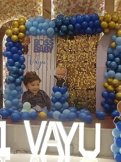 The Boss Baby theme is a huge hit for first birthdays. Here is a complete stage setup for Vayu's party in Noida, featuring a custom backdrop with his photo, character cutouts, and a blue and gold balloon arch, all tied together with large "1 VAYU" marquee letters.