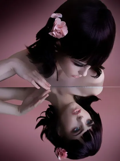 A creative beauty portrait showing a model with a sleek black bob and a pink flower in her hair, gazing at her own reflection in a mirror. This image showcases my technical skill with lighting and composition for artistic and conceptual photoshoots.