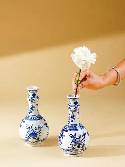A hand places a white carnation into a blue and white ceramic vase. This simple, elegant action shot adds a human element and a touch of grace to the product photography.