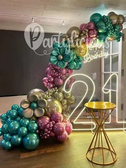 Turning 21 in style. This setup features a glowing neon '21' sign, framed by a spectacular, multi-colored metallic balloon garland with floral accents. It's a modern, edgy, and totally photo-worthy look.