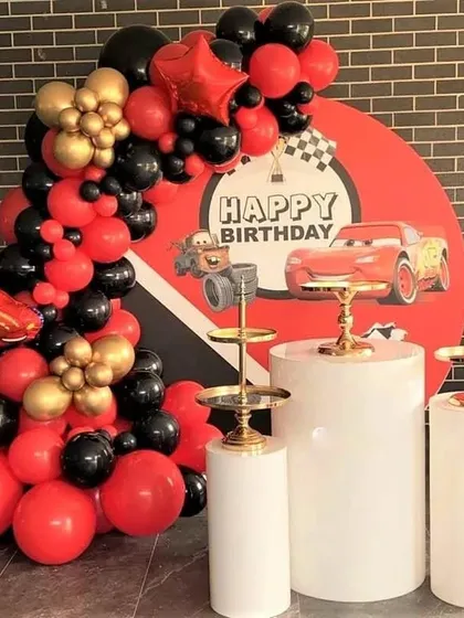A Disney Cars themed birthday backdrop with a balloon arch in red and black. The setup includes plinth stands for cake and desserts.