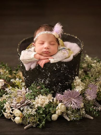 A wider view of the classic bucket pose, showing the baby surrounded by a delicate wreath of white and purple flowers on a dark wooden floor.