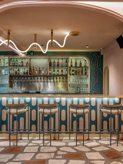 The bar at Hangover features a playful design with a custom-patterned counter, teal wallpaper, and a unique, wave-like neon light fixture. The crazy-paving terrazzo floor adds another layer of texture and color to this eclectic space.