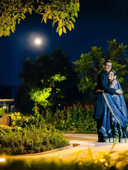 A romantic nighttime portrait of a couple under the full moon. The ambient light from the moon and the garden creates a serene and magical atmosphere.