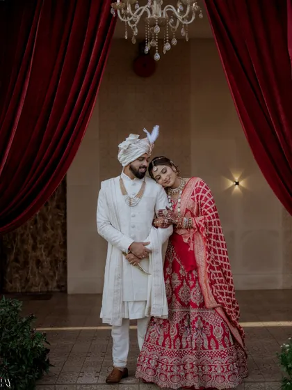 A regal and romantic portrait of the newlywed couple. The bride rests her head on the groom's shoulder, framed by dramatic red curtains that add a touch of theatrical elegance.