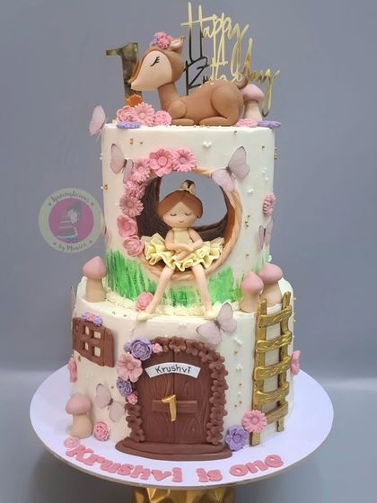 A whimsical two-tier fairyland cake for a first birthday, with a ballerina figurine sitting inside a cutout, surrounded by butterflies and mushrooms.