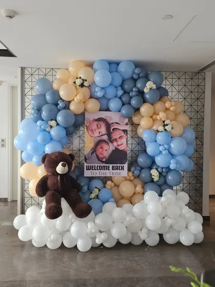 Make your welcome home celebration deeply personal with a custom photo display. This beautiful balloon arrangement in shades of blue, peach, and white frames a personalized poster featuring the family's photos, creating a heartwarming focal point to welcome the new baby.