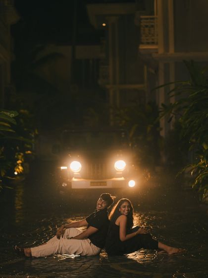 A fun and intimate night shot of a couple sitting back-to-back on a wet pavement, illuminated by car headlights. This image showcases a playful yet dramatic pre-wedding photography style.
