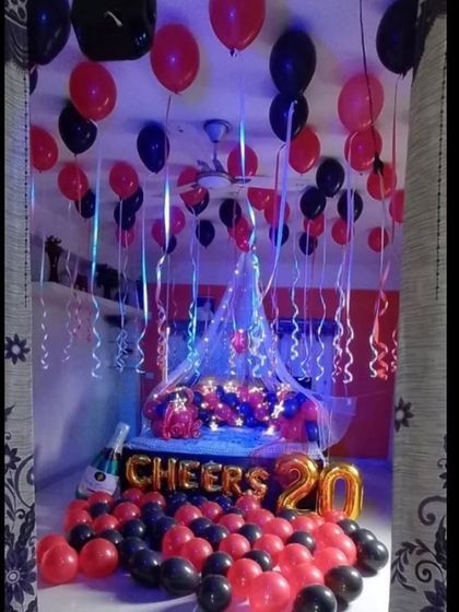 A bold and stylish anniversary or birthday setup in red and black. The room features a bed canopy with fairy lights, 'CHEERS' foil balloons, and a floor covered in balloons for a fun, party atmosphere.
