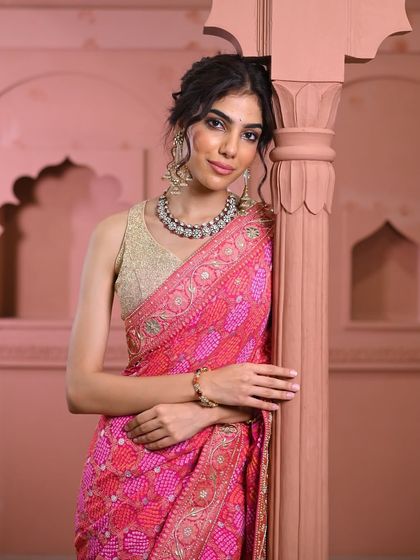 A model leans gracefully, showcasing a pink bandhani saree with a detailed gold border, paired with a simple gold blouse and traditional jewelry.