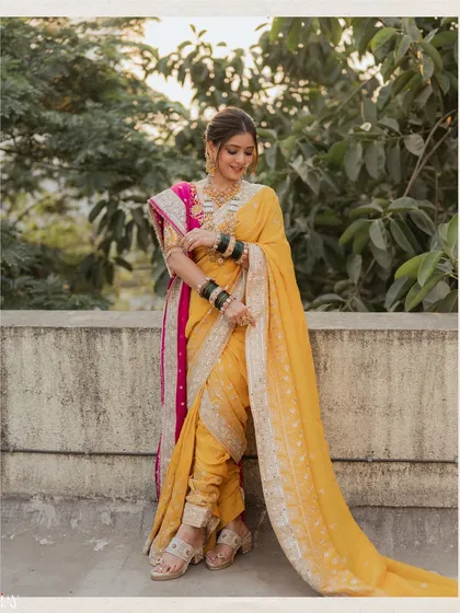 A full-length portrait of the bride in a stunning yellow nauvari saree, styled traditionally. The photograph is taken against a simple background to emphasize the intricate details of her attire and jewelry.