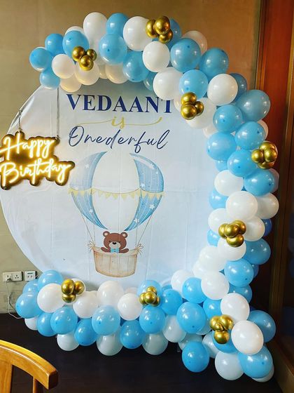 A "Onederful" hot air balloon theme for a first birthday. The custom backdrop is surrounded by a blue, white, and gold balloon arch, with a "Happy Birthday" neon sign on the side.