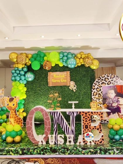A wide shot of a jungle-themed first birthday. This setup includes a green wall backdrop, a 'ONE' marquee table, and multiple animal character cutouts.
