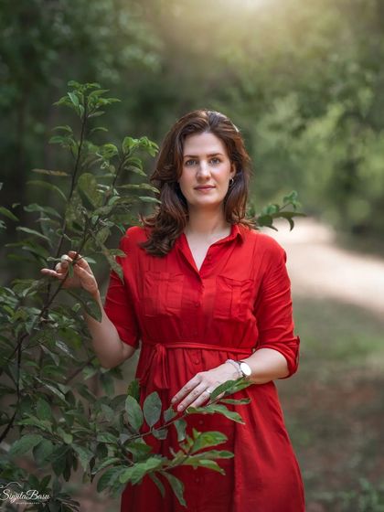 A woman in a vibrant red dress stands among the trees, the color creating a striking contrast with the green foliage. This outdoor portrait uses natural light to create a soft, painterly effect, highlighting her presence in the serene landscape.