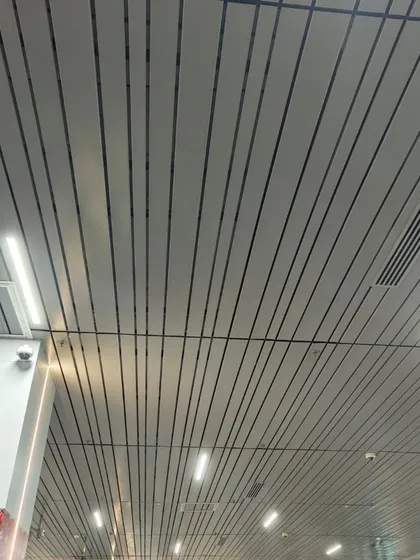 A modern, linear metal ceiling in a commercial space. The sleek white panels with black grooves integrate lighting and air conditioning vents for a clean, minimalist finish.