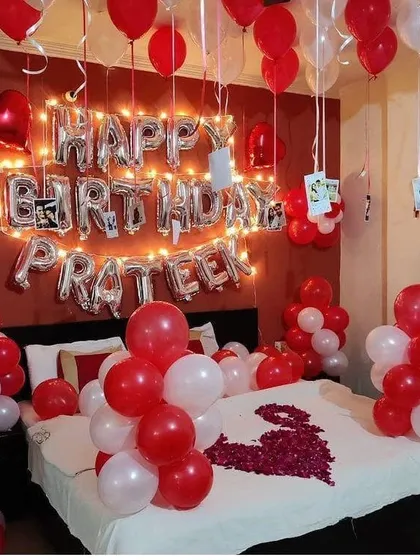 A personalized romantic birthday room decoration with the name "PRATEEK" in foil balloons, fairy lights, and photos hanging from the ceiling.