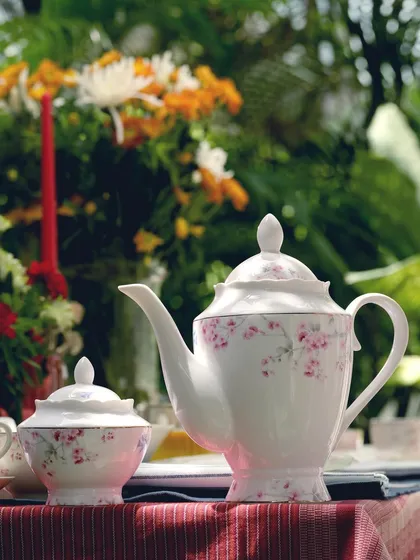 A close-up of a delicate pink floral teapot from Vigneto. The outdoor setting with blurred flowers in the background enhances the product's romantic and classic charm.