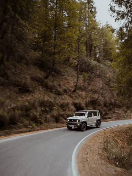 A white Suzuki Jimny is captured in a smooth panning shot as it rounds a bend on a forest road. The motion blur in the background conveys speed and agility, making for a compelling action shot.