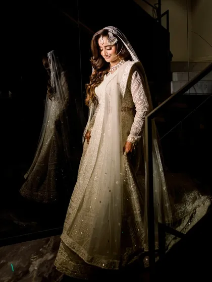 A full-length portrait of a bride in a magnificent white lehenga, her reflection captured in the glossy wall beside her. The dramatic lighting highlights the intricate details of her outfit.