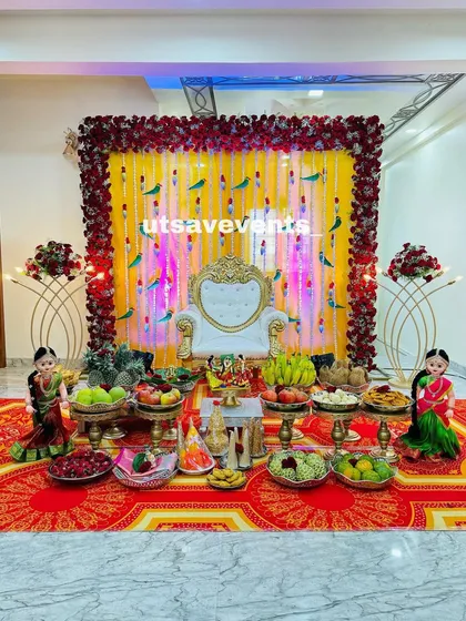 This vibrant baby shower stage uses a yellow backdrop with parrot hangings and a red rose border. The white throne and traditional dolls complete this colorful setup.