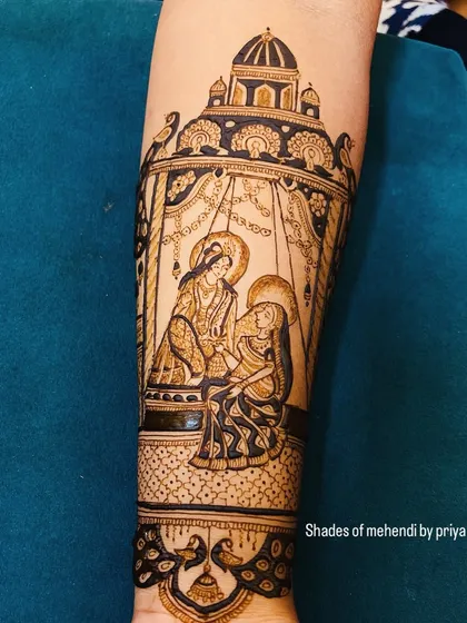 A close-up of a Radha-Krishna scene on the forearm, depicted sitting on a swing (jhula). This is a classic and romantic portrayal, perfect for bridal mehendi.