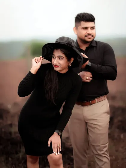 A stylish pre-wedding portrait of a couple in a field, her black dress and hat adding a touch of chic.