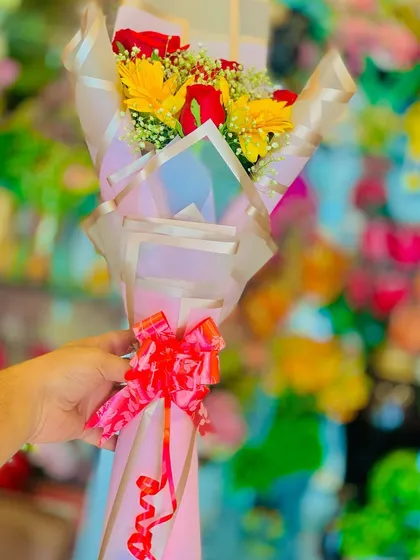 A small, hand-held bouquet of red roses and yellow gerbera daisies. The iridescent wrapping paper and red bow make it a cheerful and affordable gift.
