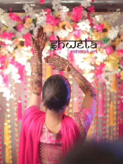 A candid shot of the bride with her hands raised, showing the full length of her stunning arm mehendi. The floral background beautifully complements the intricate henna art.