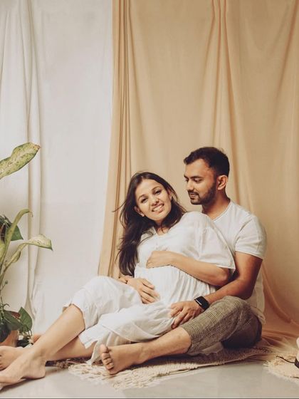 A relaxed, bohemian-style maternity shoot with the couple dressed in all white. The neutral-toned fabric backdrop and potted plant add a natural, earthy element to the portrait.